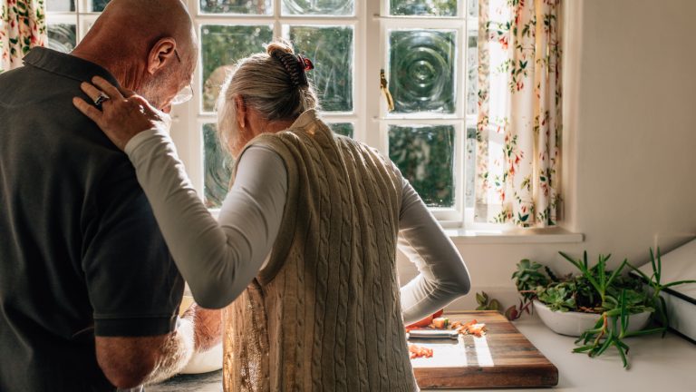 Rear view of a senior couple standing together in kitchen cooking food. Elderly woman watching her husband cook food standing in kitchen.